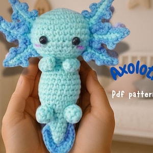 Peut inclure: Un jouet en crochet axolotl amigurumi bleu et bleu clair. L'axolotl tient ses bras devant lui et a une petite queue. Le jouet est tenu dans les mains d'une personne. Le texte "Axolotl Pdf pattern" est en arrière-plan.