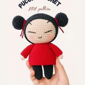 May include: A crocheted doll with a red dress, black pants, and black hair buns. The doll has a smiling face with rosy cheeks. The text "PUCCA CROCHET PDF pattern" is arched above the doll.