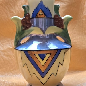 May include: A ceramic vase featuring a cream-colored base with a prominent blue and orange geometric triangle pattern. The vase has two side handles adorned with brown textured accents and green leaves, finished with a blue base and rim.