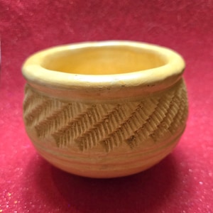 May include: A small, round, light yellow ceramic bowl. The exterior features a textured, carved pattern of diagonal lines. The rim is smooth, and the interior is a slightly darker yellow. The bowl is likely handmade and has a rustic appearance.