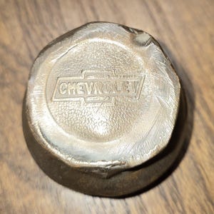 May include: A vintage, metallic Chevrolet hubcap center. The hubcap is a dull silver color with the word "CHEVROLET" embossed in a rectangular frame. The hubcap has a slightly worn appearance, suggesting age and use.