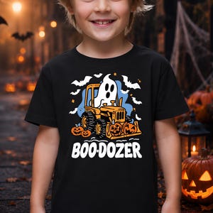 Pode incluir: Uma t-shirt preta de manga curta com um gráfico de um fantasma branco a conduzir um bulldozer cor de laranja. O design inclui abóboras cor de laranja, morcegos brancos e o texto "BOO-DOZER" em letras maiúsculas brancas sob a ilustração.