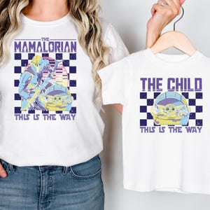 A woman wearing a white t-shirt with a graphic design featuring a character from the Star Wars series, The Mandalorian, and a child wearing a smaller t-shirt with the same design.