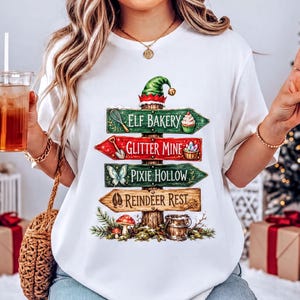 Pode incluir: Uma t-shirt branca de manga curta com um gráfico de uma placa de sinalização de madeira encimada por um chapéu de elfo. As placas dizem Elf Bakery, Glitter Mine, Pixie Hollow e Reindeer Rest, decoradas com ícones como um batedor e cupcake.