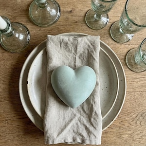 Puede incluir: Un objeto en forma de corazón de color verde salvia claro descansa sobre una servilleta de lino beige doblada, colocada sobre dos platos de cerámica de color blanco roto con motas. Rodean los platos dos candelabros y tres copas de cristal.