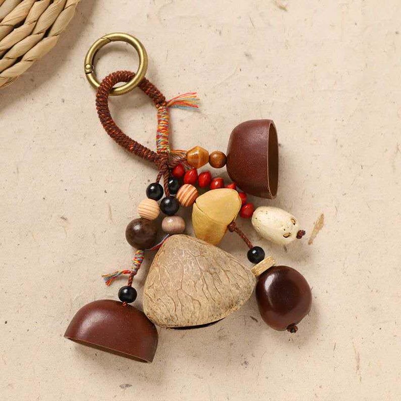 May include: A handcrafted keychain featuring a variety of natural elements. It includes brown, beige, and cream-colored beads, wooden shapes, and a brass-colored ring. The keychain is made with a woven brown cord and colorful accents.