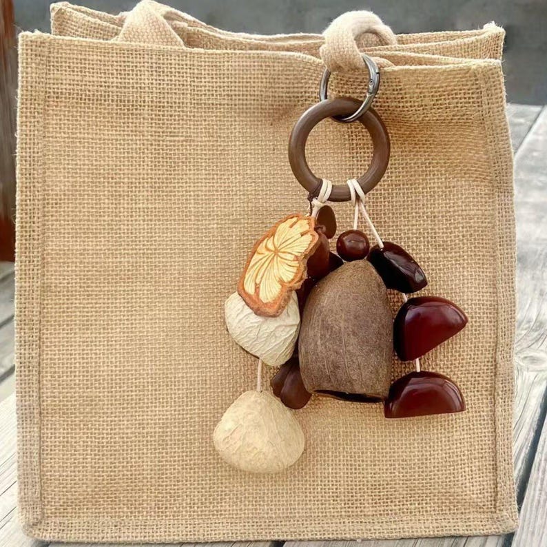 Puede incluir: Un bolso tote de yute beige con un adorno decorativo. El adorno presenta varios elementos naturales, incluyendo una pieza de madera tallada, una vaina de color claro y varias formas de semillas de color marr&oacute;n oscuro y rojizo. El bolso tiene un dise&ntilde;o sencillo.