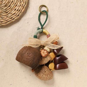 May include: A decorative keychain featuring a ring with a green cord loop and a gold-colored ring. The keychain includes a jute bow, a brown coconut shell, dried fruit slices, and dark brown, heart-shaped objects. The background is a light beige.