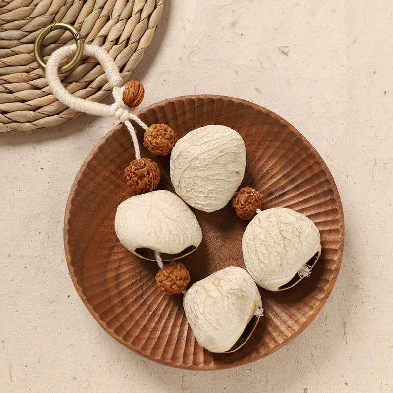 May include: A wooden bowl with a carved design holds four white, textured seed pods. Small, round, brown beads are interspersed with the pods. A woven coaster and a keychain with a white cord and gold ring are in the background.