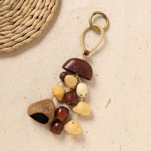 May include: A handcrafted keychain featuring a collection of natural, organic elements. The keychain includes a dark brown, half-dome shape, various seed pods, and small, rounded wooden beads. A brass ring and beige cord complete the design.