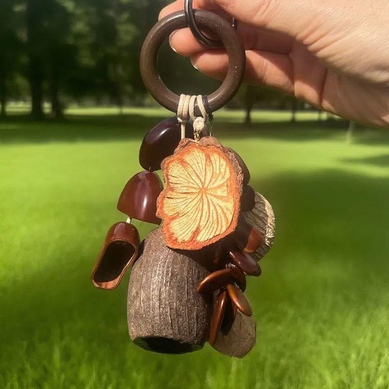 Puede incluir: Un llavero de madera hecho a mano con un anillo de madera marr&oacute;n oscuro y una colecci&oacute;n de elementos naturales. Los elementos incluyen una rodaja de madera, varias semillas y peque&ntilde;as formas de madera, todo conectado con hilo.