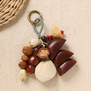 May include: A unique keychain featuring a collection of natural elements. It includes various nuts, seeds, and wooden beads in shades of brown, beige, and cream. The keychain has a gold-colored clasp and a teal cord loop.