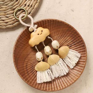May include: A wooden cloud-shaped keychain with a smiling face, connected to a beige rope and gold ring. The keychain has three tassels with beige beads and white fringe, resting on a wooden plate.