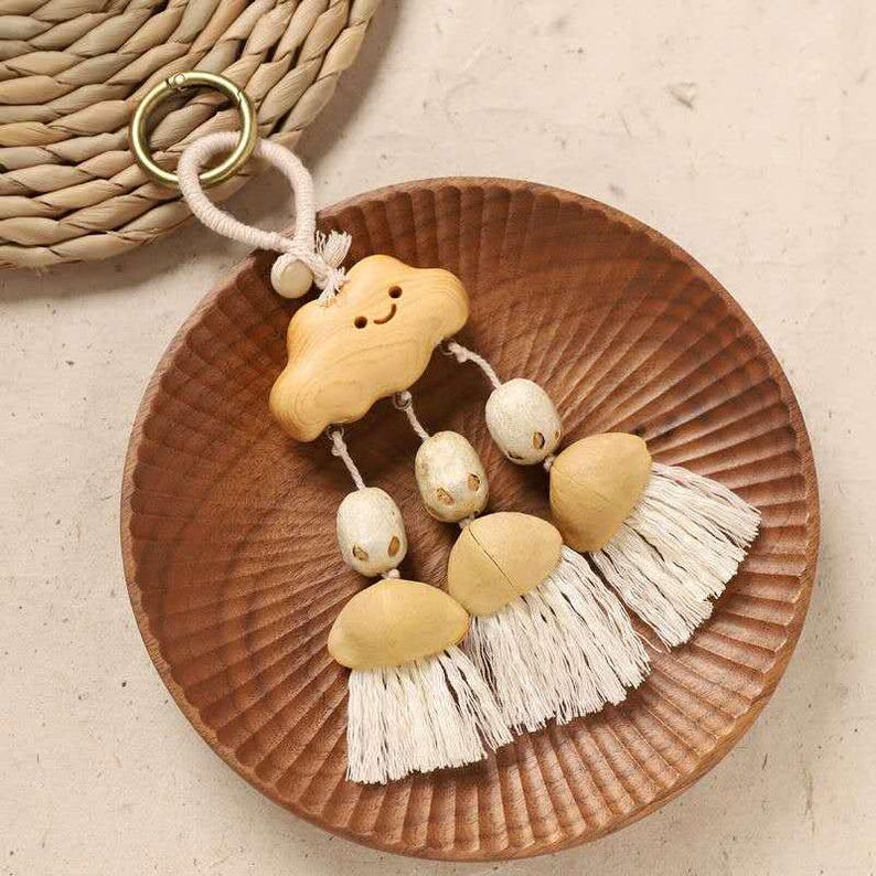 May include: A wooden cloud-shaped keychain with a smiling face, connected to a beige rope and gold ring. The keychain has three tassels with beige beads and white fringe, resting on a wooden plate.