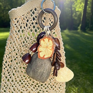 Puede incluir: Un llavero hecho a mano con un anillo de madera y varios elementos naturales. El dije incluye una rodaja de madera tallada con detalles en naranja, vainas de semillas de color marr&oacute;n oscuro y una piedra gris. El llavero est&aacute; sujeto a una bolsa de crochet beige.
