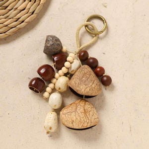 May include: A handmade keychain featuring natural elements. The keychain includes brown, beige, and cream-colored beads, seed pods, and wooden elements. A gold-colored metal ring and a woven cord complete the design. The keychain is a unique, nature-inspired accessory.