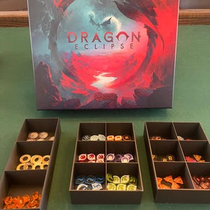 May include: A board game box with the title "Dragon Eclipse" and a dragon illustration. The box is open and shows various colored game pieces in separate compartments.