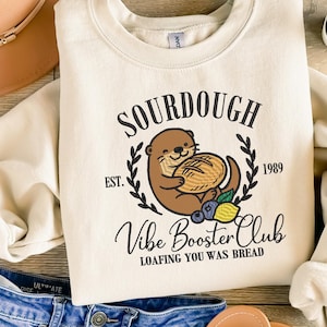 Könnte beinhalten: Cremefarbenes Sweatshirt mit einem Aufdruck, der einen Otter mit Brot zeigt, umgeben von einem Lorbeerkranz. Der Text lautet "SOURDOUGH", "Vibe Booster Club" und "Loafing You Was Bread". Das Design enthält auch das Jahr "1989" und Illustrationen von Blaubeeren und einer Zitrone.