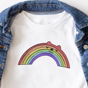 May include: White t-shirt with a rainbow design and a bear face. The rainbow has red, orange, yellow, green, blue, and purple stripes. The shirt is shown on a denim jacket.