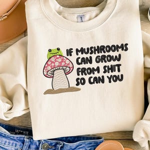 May include: Cream-coloured sweatshirt featuring the embroidered text "IF MUSHROOMS CAN GROW FROM SHIT SO CAN YOU." The design includes a cartoon mushroom with a pink and white spotted cap and a green frog. The sweatshirt is paired with blue jeans.