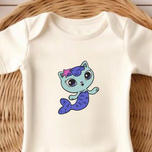 Puede incluir: Body de bebé color crema con un diseño de gato sirena de dibujos animados. La ilustración muestra un gato azul con una cola morada y una estrella de mar rosa. El body tiene mangas cortas y cuello redondo.