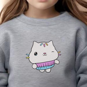 May include: A grey sweatshirt featuring a white cat wearing a pink and blue jumper. The cat has colourful stripes on its head and is winking.