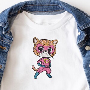 May include: White t-shirt with an embroidered image of a cartoon cat wearing a pink superhero costume. The cat has orange and brown stripes and a pink mask with a yellow heart.