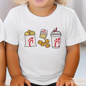 May include: White t-shirt with embroidered images of a bag of nuggets, sauce, and a milkshake. The bag and milkshake have a red heart and a stylized "C" design. The shirt is a children's size.