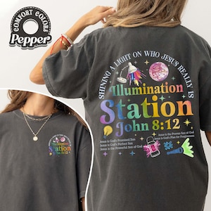 May include: Dark gray t-shirt with a colorful graphic on the back that reads "Illumination Station John 8:12." The design includes a light, a disco ball, and other illustrations. The shirt also has the Comfort Colors and Pepper logos.