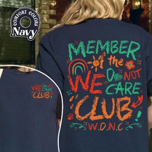 May include: Navy blue t-shirt with the text "MEMBER of the WE DO NOT CARE CLUB" in green, orange, and red. The design includes a rainbow, sun, and floral elements. The left side of the shirt has the text "WE DO NOT CARE CLUB".