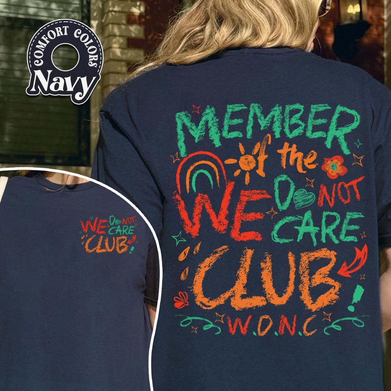 May include: Navy blue t-shirt with the text "MEMBER of the WE DO NOT CARE CLUB" in green, orange, and red. The design includes a rainbow, sun, and floral elements. The left side of the shirt has the text "WE DO NOT CARE CLUB".