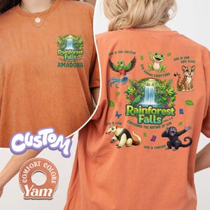 May include: Orange t-shirt with a rainforest graphic on the front and back. The front features text that reads "Rainforest Falls Amadora". The back has a waterfall scene with various animals and text that reads "Rainforest Falls Exploring the Nature of God".