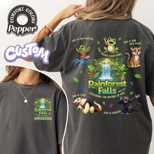 May include: Dark grey t-shirt with a vibrant rainforest design, including the text "Rainforest Falls" and illustrations of animals. The shirt also features the text "Exploring the Nature of God" and "Amadora".
