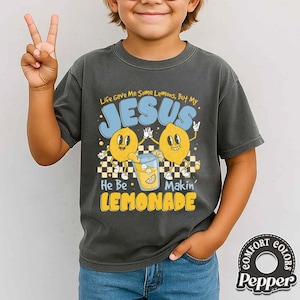 May include: Dark gray t-shirt with a graphic featuring two cartoon lemons, a glass of lemonade, and the text "Life Gave Me Some Lemons, But My JESUS He Be Makin' LEMONADE". The shirt also has the Comfort Colors Pepper logo.