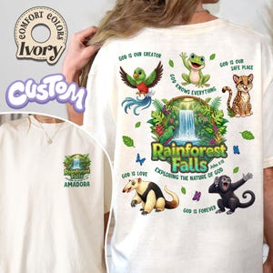 May include: Ivory t-shirt featuring a rainforest-themed design with a waterfall, animals, and the text "Rainforest Falls" and "Exploring the Nature of God". The design also includes the phrases "God is our Creator" and "God is Love".