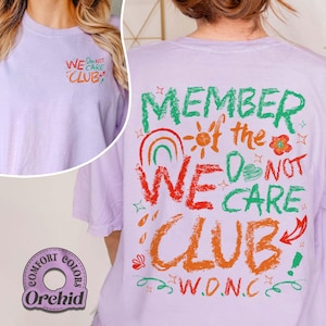May include: A light purple t-shirt with the text "WE DO NOT CARE CLUB" in colorful lettering. The back of the shirt features the phrase "MEMBER of the WE DO NOT CARE CLUB" with a rainbow, sun, and floral graphics. The shirt is labeled "Comfort Colors Orchid".