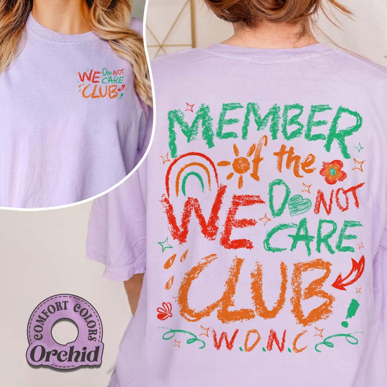 May include: A light purple t-shirt with the text "WE DO NOT CARE CLUB" in colorful lettering. The back of the shirt features the phrase "MEMBER of the WE DO NOT CARE CLUB" with a rainbow, sun, and floral graphics. The shirt is labeled "Comfort Colors Orchid".