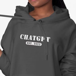 Peut inclure: Un sweat à capuche gris foncé avec le texte blanc "CHATGPT EST. 2022". Le sweat à capuche a une poche avant et une capuche à cordon. La personne porte un bonnet bordeaux et un jean bleu.