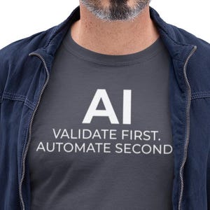 Könnte beinhalten: Ein Mann trägt ein dunkelgraues T-Shirt mit dem weißen Text "AI VALIDATE FIRST. AUTOMATE SECOND." Er trägt auch eine marineblaue Jacke, blaue Jeans und eine Brille. Er hält einen silbernen Laptop.
