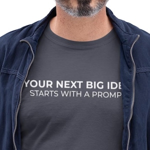Könnte beinhalten: Ein Mann trägt ein dunkelgraues T-Shirt mit dem Text "YOUR NEXT BIG IDEA STARTS WITH A PROMPT". Er trägt auch eine marineblaue Jacke, blaue Jeans und eine Brille und hält einen silbernen Laptop.