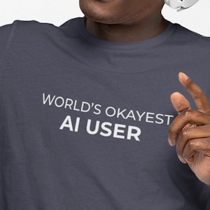 Könnte beinhalten: Eine Person mit blauer Mütze, Brille und weißen Kopfhörern, die ein rot-schwarzes Smartphone hält. Die Person trägt ein dunkelgraues T-Shirt mit dem weißen Text "WORLD'S OKAYEST AI USER".