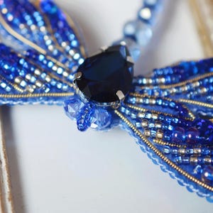 May include: A handcrafted, beaded dragonfly brooch in shades of blue, with a large, dark blue gemstone at its center. The wings are intricately detailed with small beads and gold accents, creating a shimmering effect. The brooch is set against a decorative, off-white frame.