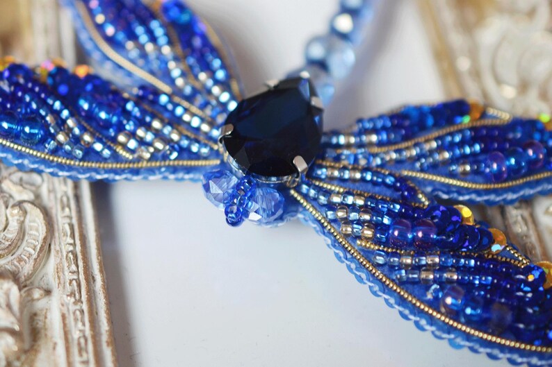 May include: A handcrafted, beaded dragonfly brooch in shades of blue, with a large, dark blue gemstone at its center. The wings are intricately detailed with small beads and gold accents, creating a shimmering effect. The brooch is set against a decorative, off-white frame.