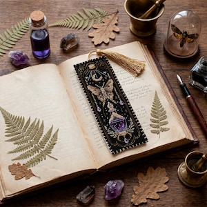 Peut inclure: Un marque-page rectangulaire noir orné d'une broderie dorée et violette représentant un papillon de nuit, un croissant de lune, un soleil et une fiole. Le marque-page est doté d'un pompon doré et posé sur un livre ouvert avec des feuilles séchées.
