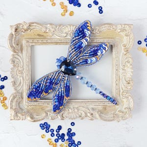 May include: A handcrafted, beaded dragonfly brooch in shades of blue and gold, resting on a vintage-style, ornate picture frame. The dragonfly's wings are detailed with sequins and beads. Scattered sequins in matching colors surround the frame.