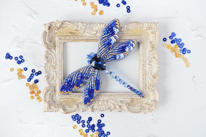 May include: A handcrafted, beaded dragonfly brooch in shades of blue and gold, resting on a vintage-style, ornate picture frame. The dragonfly's wings are detailed with sequins and beads. Scattered sequins in matching colors surround the frame.