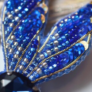 May include: Close-up of a blue beaded brooch. The brooch features a dark blue gemstone at the base and two wings adorned with blue sequins, beads, and gold accents. The wings have a fan-like design.