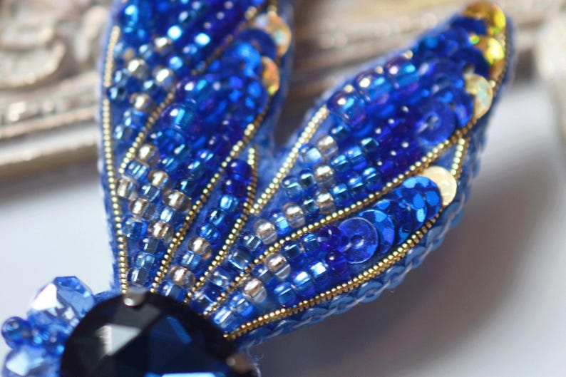 May include: Close-up of a blue beaded brooch. The brooch features a dark blue gemstone at the base and two wings adorned with blue sequins, beads, and gold accents. The wings have a fan-like design.