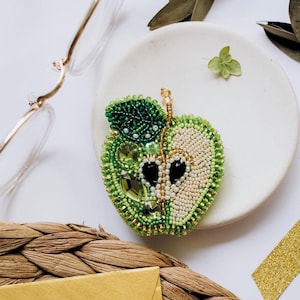 Op de afbeelding: Een handgemaakte, met kralen versierde appelbroche. De broche is groen en wit, met een gedetailleerd ontwerp van een doorgesneden appel. De broche heeft een groen kralenblad en is versierd met diverse groene kralen en kristallen. De broche ligt op een wit bord.