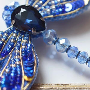 May include: A handcrafted, blue dragonfly brooch. The wings are embellished with sequins and beads in shades of blue and gold. A large, faceted, dark blue gem is at the center. A beaded strand extends from the dragonfly's body.